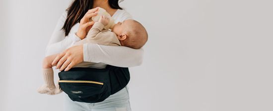 Parent feeding baby with a bottle while using the Tushbaby Hip Carrier for support during feeding time.