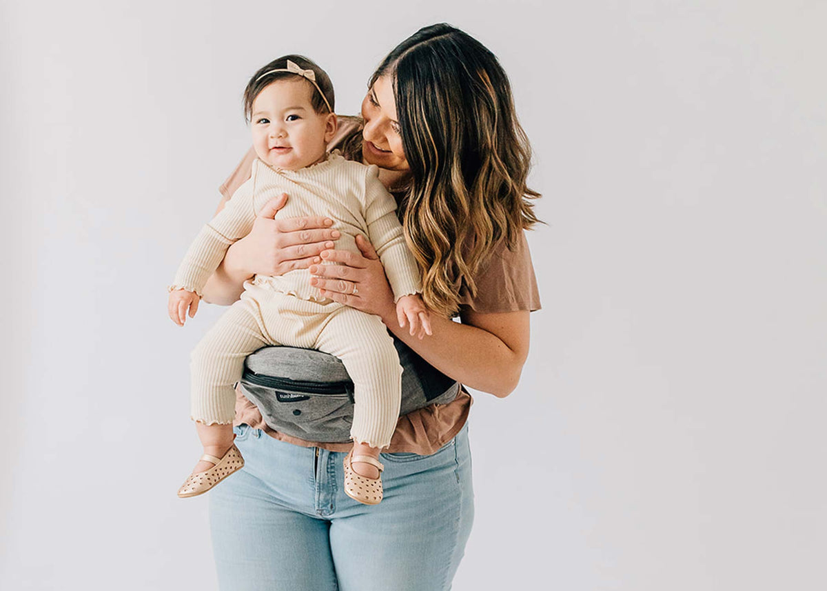 Parent smiling while holding baby in a front-facing position on a Tushbaby Hip Carrier, showing comfort and support.