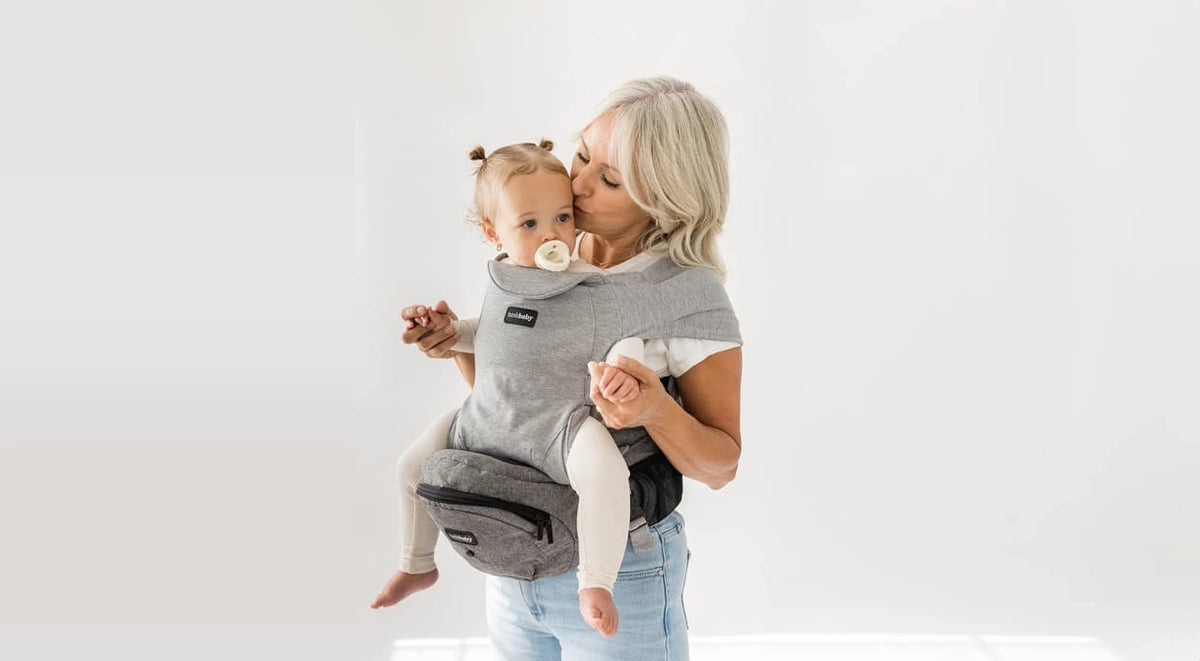 Grandmother holding and kissing baby in a gray Tushbaby hip carrier with pacifier, showing close bonding and hands-free support.