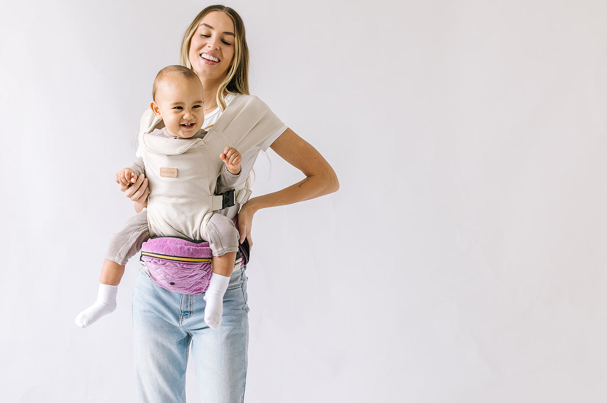 Smiling baby carried in a beige hands-free attachment, sitting on a purple Tushbaby hip carrier held by a parent.
