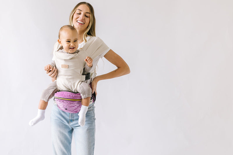 Smiling baby carried in a beige hands-free attachment, sitting on a purple Tushbaby hip carrier held by a parent.
