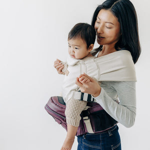 Adult carrying a baby facing outward in a Cream Snug Attachment on a Vegan Leather Mulberry Tushbaby Hip Carrier against a white background