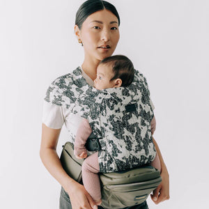 Adult carrying a baby facing inward in an Abstract Snug Attachment connected to a Vegan Leather Olive Tushbaby Hip Carrier against a white background