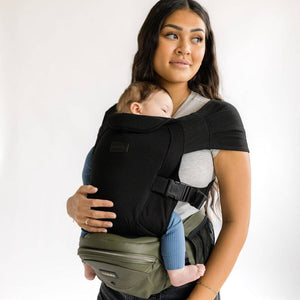 Adult carrying an infant in the Black Snug Attachment connected to a Vegan Leather Olive Tushbaby Hip Carrier against a white background