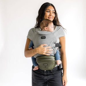 Adult carrying an infant in the Grey Snug Attachment connected to a Vegan Leather Olive Tushbaby Hip Carrier against a white background