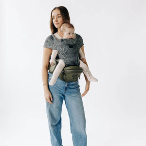 Adult carrying a baby facing outward in a Jaguar Snug Attachment connected to a Vegan Leather Olive Tushbaby Hip Carrier against a white background