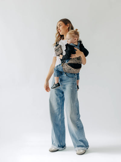 Full-length view of an adult carrying a child in the Cheetah Tushbaby Liftoff Toddler Sling against a white background