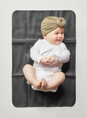 Smiling baby lying on unfolded The Changing Pad in Black, wearing a white bodysuit and knotted headwrap